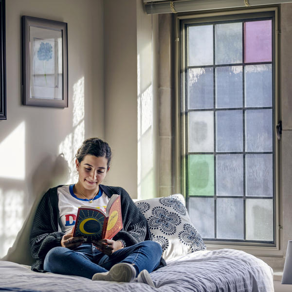 A young woman sits on her bed reading by the light of a stained glass window.
