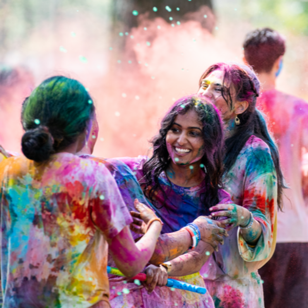 A young woman with an infectious smile is covered in brightly colored chalk. Behind her the Holi festivities continue.